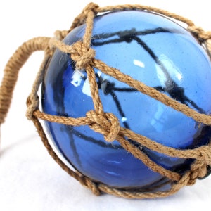 Op de afbeelding: Een blauwe glazen vissersspeer met een natuurlijk touwnet. De drijver heeft een lus van touw om op te hangen.