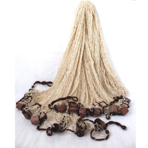 Könnte beinhalten: Ein großes, cremefarbenes Fischernetz mit braunen Holzgewichten. Das Netz ist gefaltet und über eine weiße Oberfläche gelegt.