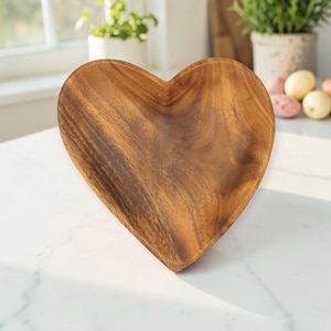 Può includere: Una ciotola in legno a forma di cuore caratterizzata da una superficie liscia e lucida con venature naturali visibili in varie tonalità di marrone, dal medio allo scuro. La ciotola presenta un centro concavo e una sagoma a cuore.