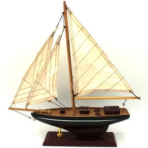 Op de afbeelding: Een modelzeilboot met een donkerbruine houten romp met een witte horizontale streep, twee gebroken witte stoffen zeilen met stikseldetails, een hoge houten mast en een rechthoekige houten voet met een goudkleurige metalen steun.