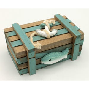 Può includere: Una scatola rettangolare in legno costruita con doghe orizzontali con una finitura usurata color verde acqua e legno naturale. Il coperchio presenta un'ancora in legno bianco avvolta da una corda beige e un pesce in legno sul davanti.