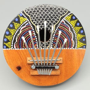 Könnte beinhalten: Eine runde Holzkalimba mit einem schwarz, weiß, gelb, rot und blau bemalten Design. Die Kalimba hat acht Metallzinken und ein Schallloch in der Mitte.