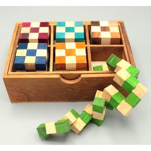 Könnte beinhalten: Ein Holzpuzzleset mit sechs farbigen Würfelpuzzles in einer Holzkiste. Die Würfel haben Schachbrettmuster in verschiedenen Farben, darunter Blau, Orange und Lila. Ein separates grünes und beiges Schlangenpuzzle ist ebenfalls enthalten.