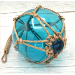 Könnte beinhalten: Ein blauer Glas-Schwimmer mit einem Netz aus Naturseil. Der Schwimmer hat eine kleine blaue Glasperle am Boden.