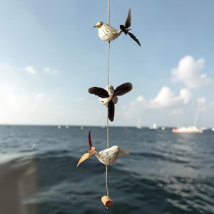 Könnte beinhalten: Ein vertikales Hängemobil mit drei kleinen Vogelfiguren, die an einer dünnen Schnur befestigt sind. Jeder Vogel hat einen weißen Körper mit dunkelbraunen Sprenkeln und Flügel aus dunkelbraunen Federn. Ein kleiner Holzzylinder hängt ganz unten.