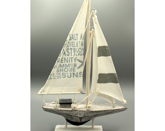 Puede incluir: Un velero de madera blanco con una vela blanca con rayas grises y texto azul. El texto en la vela dice "Salt Air, Shovel 6 Pa, East Buoy, Serenity, Summer Shore, 12 Seasons". El velero está en un soporte de madera blanco.