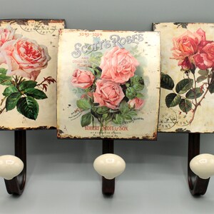 Könnte beinhalten: Drei Vintage-Metallhaken mit Porzellanknöpfen, jeder mit einem anderen Blumendesign mit rosa Rosen und grünen Blättern. Der mittlere Haken trägt den Text "Scott's Roses" und "Robert Scott & Son".