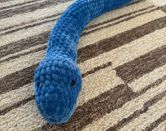 May include: A blue crocheted snake toy with black eyes. The toy is lying on a striped rug.