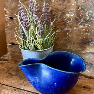 May include: A cobalt blue ceramic bowl with a unique, asymmetrical shape. The bowl sits on a weathered wooden surface, with a small metal pot of lavender in the background. The bowl's glossy finish reflects the light, highlighting its curves.