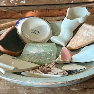 May include: A collection of antique pottery shards in a distressed, oval-shaped, light blue bowl. The fragments include pieces of white, brown, pink, and patterned ceramics, with a small white ceramic leg and a piece marked "JAPAN".