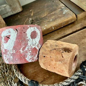 May include: Two cylindrical fishing net floats. One float is red with a weathered, chipped white painted number 78 on its surface. The second float is a smaller, unpainted, porous tan cylinder with a visible hole through the center.