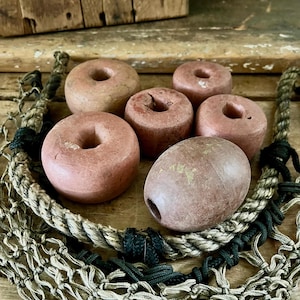 May include: A collection of six weathered, round, and oval-shaped wooden fishing net floats in shades of muted brown and reddish-pink, each featuring a central hole, arranged atop a thick, twisted beige rope and a fine-mesh fishing net.