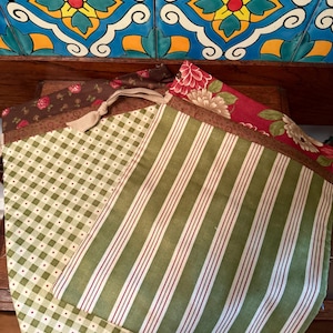 Può includere: Tre sacchetti di stoffa con coulisse e motivi diversi. Il sacchetto superiore è rosso con motivi floreali. Il sacchetto centrale è verde con strisce bianche. Il sacchetto inferiore è verde con un motivo a quadri bianchi e rossi.