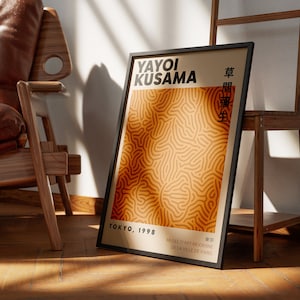 Może przedstawiać: Czarny plakat w ramie z abstrakcyjnym wzorem w kolorze pomarańczowym i brązowym. Plakat zawiera tekst "YAYOI KUSAMA" i "TOKYO, 1998". Plakat zawiera również tekst japoński i tekst "MUSEE D'ART MODERNE DE LA VILLE DE PARIS".