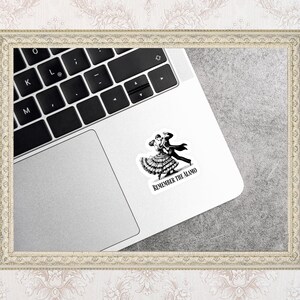 May include: A black and white sticker featuring a couple dancing, with the text "REMEMBER THE ALAMO." The sticker is placed on a silver laptop. The laptop keyboard is visible.