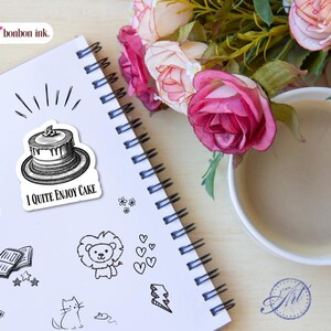May include: A spiral-bound notebook with a sticker that says "I Quite Enjoy Cake" next to other small black and white doodles. A cup of coffee and pink roses are in the background. The notebook has a white cover and black spiral binding.