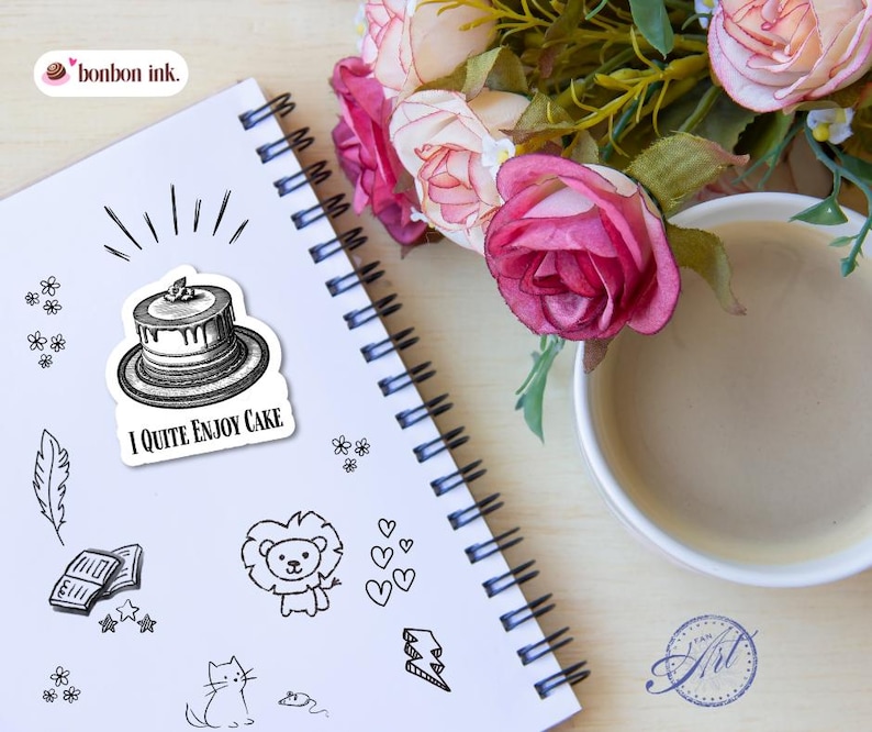 May include: A spiral-bound notebook with a sticker that says "I Quite Enjoy Cake" next to other small black and white doodles. A cup of coffee and pink roses are in the background. The notebook has a white cover and black spiral binding.