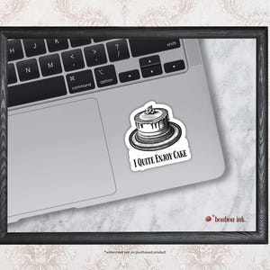 May include: A black and white sticker featuring a cake illustration with the text "I Quite Enjoy Cake" on a laptop. The sticker is on a silver laptop, with the keyboard visible. The image is framed in a black wooden frame.