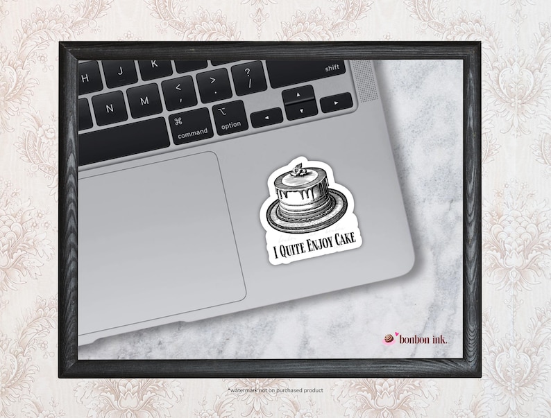 May include: A black and white sticker featuring a cake illustration with the text "I Quite Enjoy Cake" on a laptop. The sticker is on a silver laptop, with the keyboard visible. The image is framed in a black wooden frame.