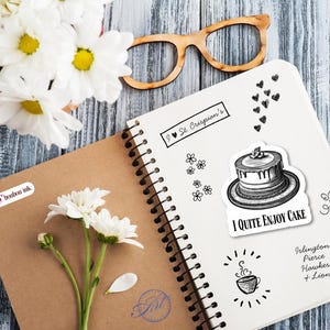 May include: An open spiral-bound notebook with a cake sticker that says "I QUITE ENJOY CAKE". A bouquet of white daisies and wooden-framed glasses are on a blue-gray wooden surface. The notebook also has the text "I St. Crispian's".