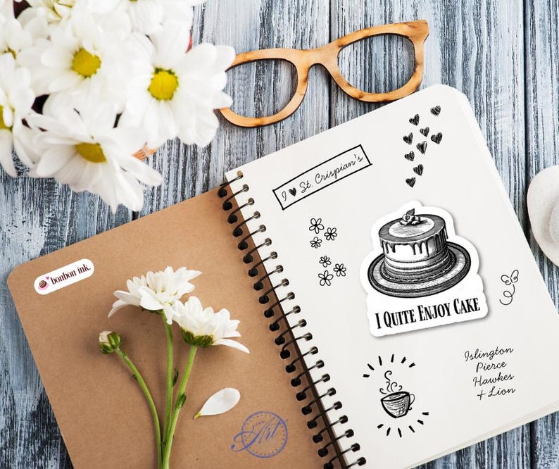 May include: An open spiral-bound notebook with a cake sticker that says "I QUITE ENJOY CAKE". A bouquet of white daisies and wooden-framed glasses are on a blue-gray wooden surface. The notebook also has the text "I St. Crispian's".