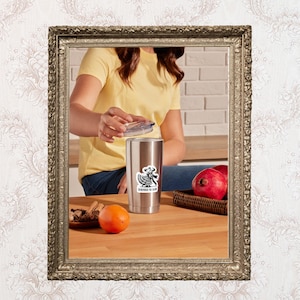 May include: A stainless steel tumbler with a black and white sticker that says "Remember the Alamo". The tumbler is on a wooden surface with an orange and a plate of cinnamon sticks. A person is holding the lid of the tumbler.
