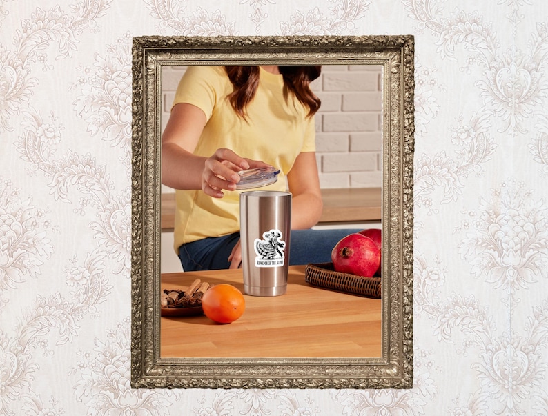 May include: A stainless steel tumbler with a black and white sticker that says "Remember the Alamo". The tumbler is on a wooden surface with an orange and a plate of cinnamon sticks. A person is holding the lid of the tumbler.