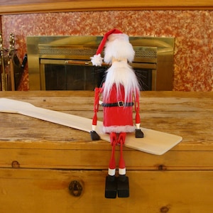 May include: A wooden Santa Claus doll with a white beard and red suit is sitting on a wooden paddle. The doll is wearing a black belt and has white accents on its boots.