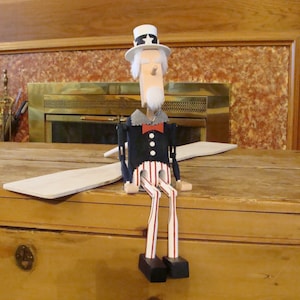 May include: A wooden Uncle Sam doll with a white hat, beard, and red bow tie. The doll is dressed in a black coat, blue trousers, and red and white striped legs. It is sitting on a wooden surface.