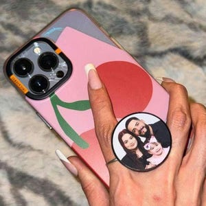 May include: A pink and green phone case with a cherry design. A black phone ring holder with a photo of a family of three is attached to the case.