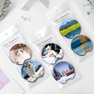 May include: Several car coasters in clear plastic packaging. The coasters feature various designs, including a cat, a mountain landscape, and a winter scene. The packaging reads "Car Coasters Handmade with love."