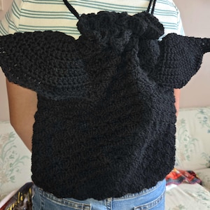 May include: A black crochet backpack with a unique design. The backpack features a drawstring closure and two wing-like extensions on the sides. The crochet work has a textured appearance, and the straps are thin and black.