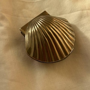 May include: A metallic, seashell-shaped compact mirror. The compact has a hinged lid and a ridged, fan-like design. The mirror is a shiny, silver color and appears to be made of metal. The background is a light tan fabric.