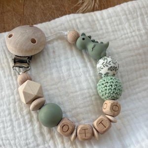 Può includere: Un fermaglio per ciuccio per bambini con una clip in legno e varie perline. La clip presenta un piccolo dinosauro verde, blocchi di lettere di legno che compongono "OTI" e altre perline decorative in tonalità verdi e legno naturale.