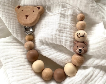 May include: A wooden baby teether with a bear head on one end and a wooden bead with the name "Noah" engraved on it. The teether is made of natural wood and silicone beads in beige and brown colors.