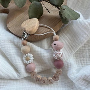 Könnte beinhalten: Ein Holz-Beißring für Babys mit einem rosa Vogel, einer weißen Margerite und Holzperlen, auf denen der Name "JOSEFA" eingraviert ist. Der Beißring ist an einem herzförmigen Holzclip befestigt.