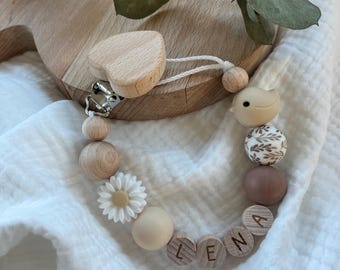 May include: A pacifier clip featuring a heart-shaped wooden attachment, a metal clasp, and a string of beads in shades of beige, brown, and white. The beads include a white daisy, a bird shape, a leaf-patterned sphere, and wooden discs spelling LENA.