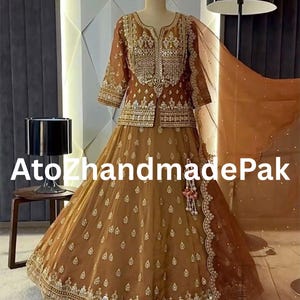 May include: A brown, embellished formal dress with a matching jacket and skirt. The outfit features intricate gold embroidery and beadwork. A sheer, brown scarf is draped over the shoulder. The text "Ato ZhandmadePak" is visible.