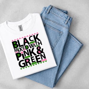 May include: A white short-sleeved t-shirt featuring the black text "BLACK MIXED WITH PINK & GREEN" with pink and green geometric accents. The shirt is paired with a pair of light blue denim jeans, showcasing a casual and coordinated clothing set.