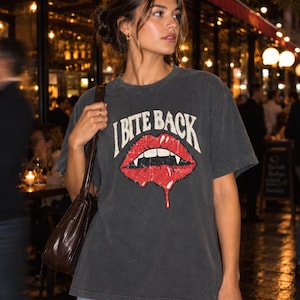 Könnte beinhalten: Ein anthrazitfarbenes Kurzarm-T-Shirt mit einer Grafik aus roten Lippen mit spitzen weißen Fangzähnen und herabtropfender roter Flüssigkeit. Der Text "I BITE BACK" ist in einer weißen, geschwungenen Schrift über den Lippen gedruckt. Rundhalsausschnitt.