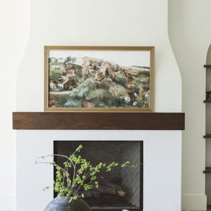 Puede incluir: Una chimenea blanca con una repisa de madera y un cuadro enmarcado de un paisaje encima. Un jarrón con ramas verdes se encuentra en una mesa de café frente a la chimenea.