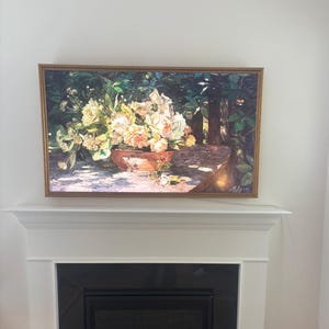 May include: A framed floral still life painting. The artwork depicts a terracotta pot overflowing with white, yellow, and peach-colored flowers. The background shows a lush green garden scene. The painting is displayed above a fireplace.