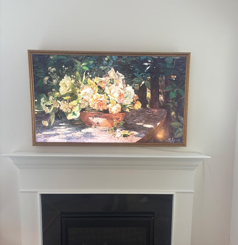 May include: A framed floral still life painting. The artwork depicts a terracotta pot overflowing with white, yellow, and peach-colored flowers. The background shows a lush green garden scene. The painting is displayed above a fireplace.