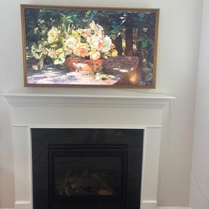 May include: A framed floral still life painting hangs above a fireplace. The painting features a terracotta bowl overflowing with white and peach-colored flowers. The frame is gold, and the fireplace is black with a white mantel.