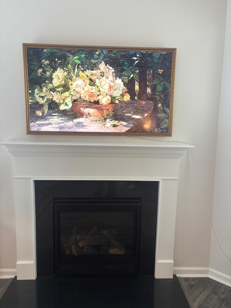 May include: A framed floral still life painting hangs above a fireplace. The painting features a terracotta bowl overflowing with white and peach-colored flowers. The frame is gold, and the fireplace is black with a white mantel.