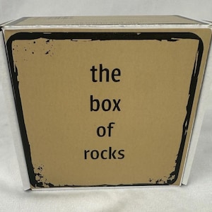 May include: A square cardboard box with a tan front panel featuring a black distressed border and the text "the box of rocks" printed in black sans-serif font. The sides of the box are white, and the top edge shows a partial barcode.