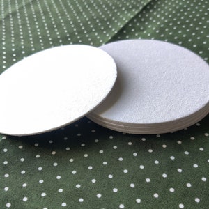 May include: A set of four white round coasters with a textured surface. The coasters are stacked on top of each other on a green background with white polka dots.