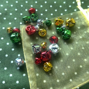 May include: A collection of small jingle bells in red, green, silver, and gold colors. The bells are scattered on a green and white polka dot fabric and a gold mesh bag.