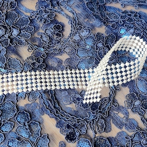 May include: A white beaded ribbon on a blue lace fabric background.