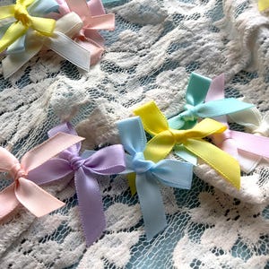 May include: A collection of pastel-colored satin bows arranged on a white lace fabric. The bows are in shades of pink, blue, yellow, and lavender.
